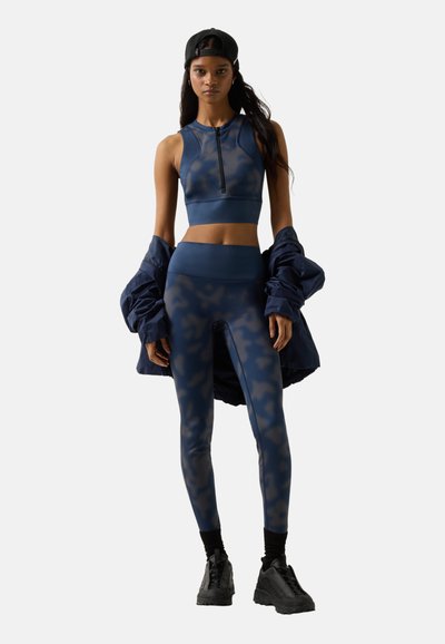 Kvinde iført navyblå camouflagemønstret crop top og leggings med sorte sneakers og kasket, holder en navyjakke hængende over armene.
