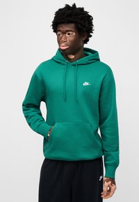 Zelena bombažna jopa s sprednjim žepom in nastavljivo kapuco z vrvico. Na levi strani prsnega koša je bel logotip Nike. Črne hlače.