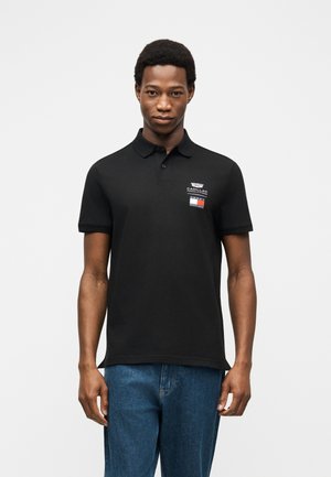 Bărbat purtând un tricou polo negru cu logo-ul Cadillac și blugi albaștri, stând în picioare pe un fundal alb simplu.