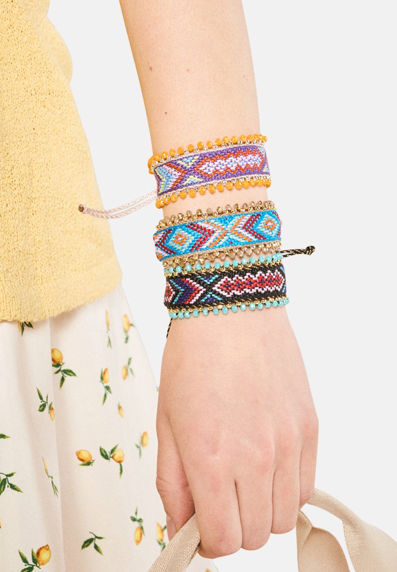 Set med fyra färgglada flätade armband med geometriska mönster i blått, lila, orange och svart, prydda med pärlor och metalliska detaljer.