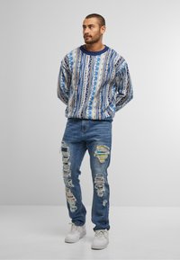 Multikleurige gebreide trui met blauwe en witte patronen, gecombineerd met versleten blauwe jeans met grote scheuren en multikleurige stoffen patches.