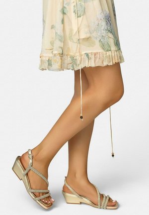Gambe con sandali dorati con cinturini e zeppa bassa abbinati a un vestito leggero con orlo arricciato a fiori, in piedi su uno sfondo semplice.