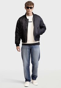 Zwarte bomberjack met een satijnen afwerking, ritsluiting en zijzakken, gedragen over een witte sweatshirt, gecombineerd met lichtblauwe jeans en witte sneakers.