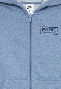 Hanorac cu fermoar bleu deschis, cu etichetă Nike și logo brodat „Paris Saint-Germain” pe piept.