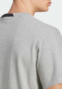 T-shirt gris clair à manches courtes en tissu doux, avec un col rond et une surface texturée discrète. Accents noirs au niveau du col arrière.