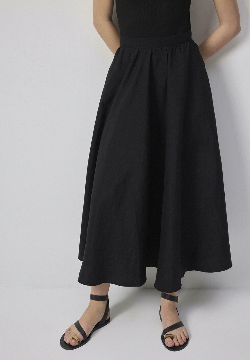 Άτομο που φοράει μακριά μαύρη φούστα και μαύρα σανδάλια ανοιχτά στα δάχτυλα με λουριά στον αστράγαλο, στέκεται μπροστά σε έναν απλό φωτεινό τοίχο.