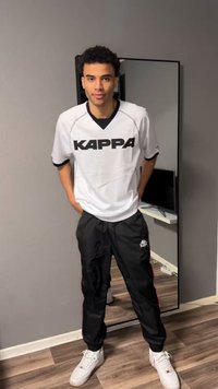 Mladý muž oblečený v bielej košeli Kappa, čiernych nohaviciach Kappa a bielych teniskách stojaci vo vnútri pred stenou a zrkadlom na celú dĺžku.