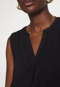 Närbild av en person som bär en ärmlös svart topp, guldkedja och stora guldringaörhängen mot en enkel bakgrund.