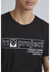 Melna kokvilnas T-kreklu ar apaļu kakla izgriezumu, kura priekšpusē ir balts teksts un ģeometrisks logo prints. Īsas piedurknes.