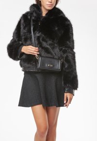Chaqueta de piel sintética negra con un ribete de cuero, combinada con una falda negra texturizada. Lleva un pequeño bolso negro con un logo dorado.