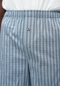 Boxer à taille élastique avec motif à carreaux gris et noir, porté avec une chemise blanche, présentant un bouton central sur la braguette.