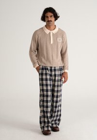 Beżowy sweter z dzianiny ze wzorem w warkocze i białym kołnierzykiem, w połączeniu z szerokimi spodniami w kratę w odcieniach granatu i kremu. Brązowe buty dopełniają stylizację.