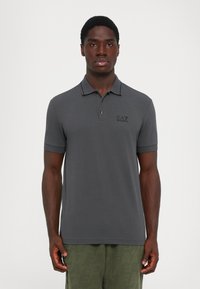 Young man standing facing forward wearing a dark gray polo shirt with logo on chest and olive green textured pants.
