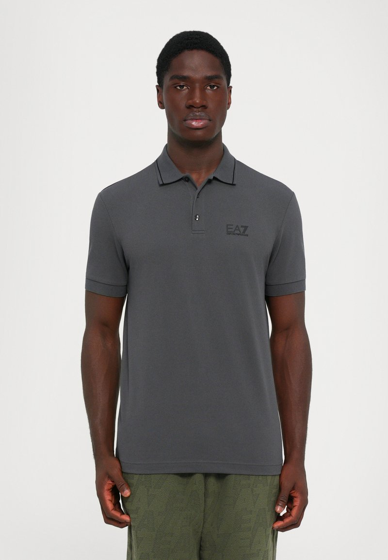Young man standing facing forward wearing a dark gray polo shirt with logo on chest and olive green textured pants.