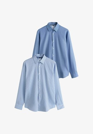 Junger Mann, der ein hellblaues Hemd, eine passende Krawatte, schwarze Hose und schwarze Lederschuhe trägt, steht mit einer Hand in der Tasche vor einer weißen Wand.