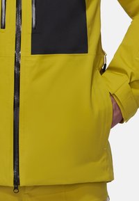 Gelbe wasserdichte Jacke mit schwarzen Akzenten, ausgestattet mit einem Frontreißverschluss, Seitentaschen und einer glatten, strukturierten Oberfläche.