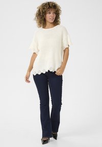Top in maglia crema con bordi ondulati, maniche corte e dettagli a occhiello. Indossato con jeans svasati blu scuro e tacchi neri a punta.