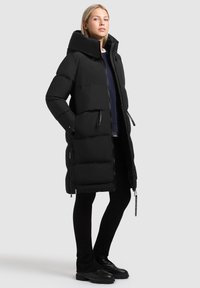 Cappotto lungo nero invernale con colletto alto, chiusura con zip e due tasche laterali. Design testurizzato e imbottito con una silhouette elegante. Indossato con pantaloni neri.