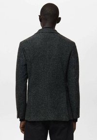 Grau strukturierter Blazer mit taillierter Passform, gekerbtem Revers und einem einzelnen Rückenschlitz. Zwei Knöpfe an den Ärmelenden; kombiniert mit dunkler Hose.