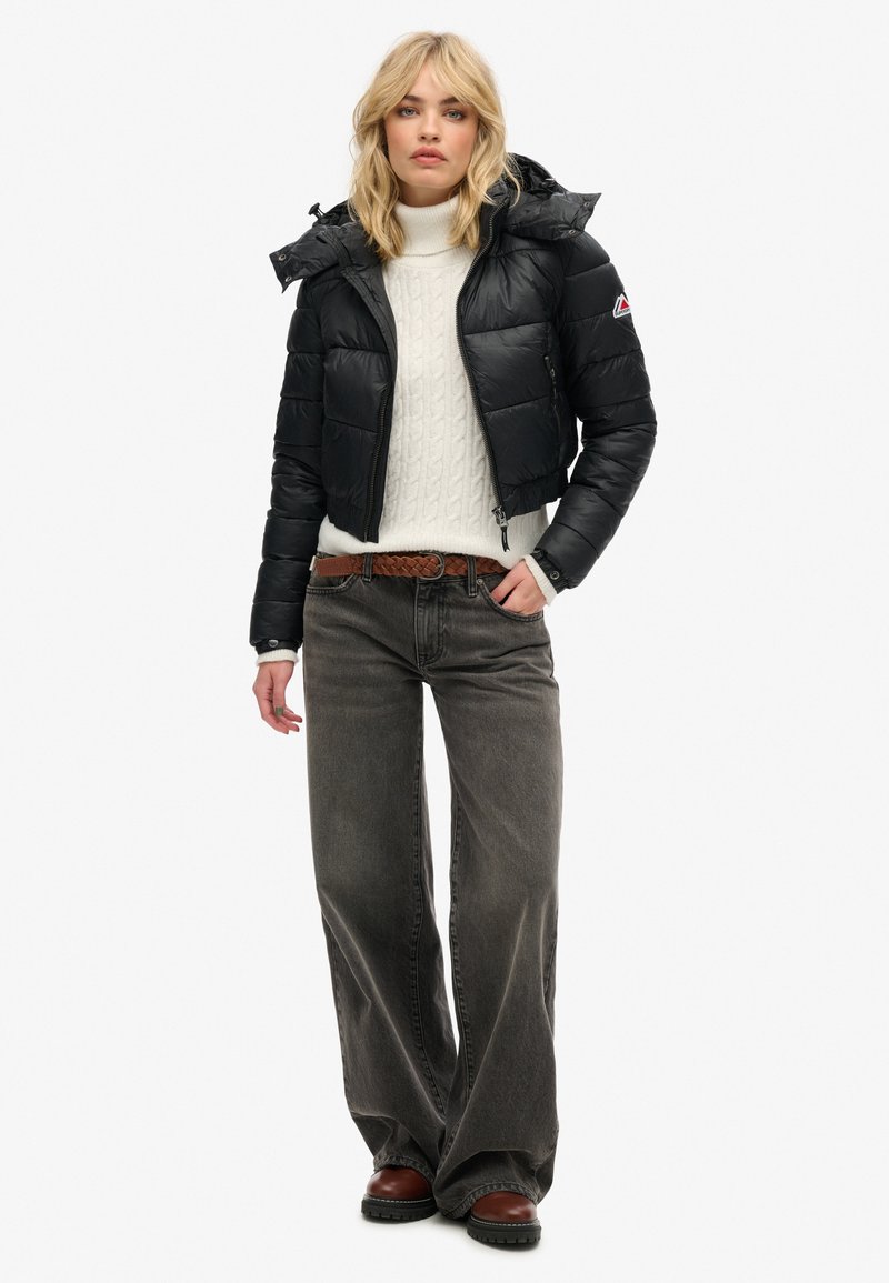 Giacca a vento nera con design corto e cappuccio, indossata sopra un maglione di lana bianco. Abbinata a jeans grigi con gamba larga e stivali marroni.