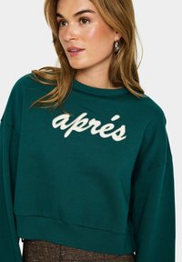 Teal cropped sweatshirt met ronde hals, met "après" in witte tekst. Gemaakt van zachte stof met drop shoulders en geribbelde zoom.