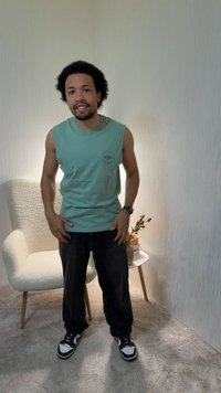 Homme portant un débardeur vert et un pantalon noir debout dans une pièce moquettée près d'un fauteuil blanc et d'une petite table ronde avec une plante et des livres.