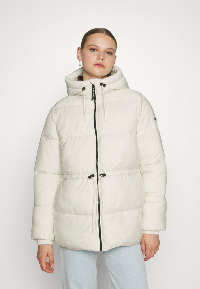 Chaqueta acolchada en crema claro con capucha, que presenta un zipper negro, cordones ajustables y una textura acolchada.