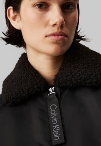 Svart jakke med en teksturert, mørkebrun fleecekrage. Har en sølvglitrende glidelås og et merkelapp med "Calvin Klein" logo i grått.