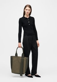 Femme portant un haut à manches longues noir et un pantalon, tenant un grand sac fourre-tout olive et vert foncé, debout devant un fond blanc.