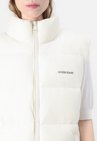 Puffer vest in gebroken wit, met een hoge kraag en een volledige rits. Zachte textuur met subtiele stiksels en een "GUESS JEANS" logo.