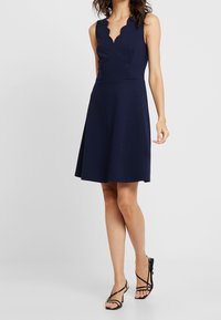 Marinblå klänning med åtsittande liv och vågig halsringning. Utsvängd kjol som sträcker sig till knäet, tillverkad av ett mjukt, stretchigt tyg.