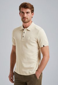 Beige Poloshirt mit kurzen Ärmeln, strukturiertem Stoff, Knopfleiste mit drei Knöpfen und einem kleinen Logo-Patch auf der linken Brusttasche.