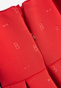 Sarkans audums ar reljefiem rakstiem, tostarp burtiem "B" un "BAKER", kā arī loku motīviem. Iekļauj rāvi, lai slēgtu. Gluda tekstūra.