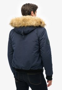 Veste matelassée marine avec capuche en fausse fourrure. Manches et taille côtelées. Présente un patch logo texturé sur la manche gauche.