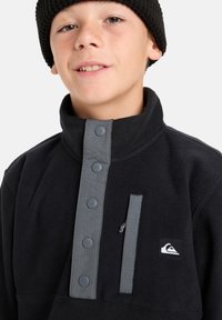 Schwarzer Fleece-Pullover mit hohem Kragen, grauer Knopfleiste, Reißverschlusstasche und Logo-Patch. Weicher Stoff und lässiges Design.