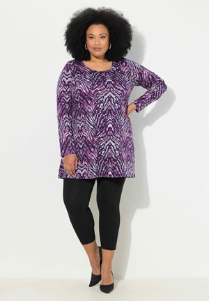 Violetti tunika, jossa on abstractti kuvio, pitkät hihat ja pyöreä pääntie. Yhdistetty mustiin legginsseihin ja teräviin mustiin korkokenkiin. Sujuva pinta.