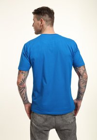 Blå kortærmet t-shirt med rund halsudskæring, lavet af bomuld, vist fra ryggen, sammen med grå bukser, som viser tatuerede arme.