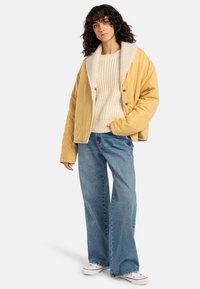 Giacca gialla imbottita con colletto foderato in shearling, indossata sopra un maglione crema e jeans larghi blu, abbinata a sneaker bianche.