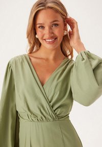 Zelené šaty s hlubokým V-výstřihem, nařaseným pasem a dlouhými balónovými rukávy; hladká látka s mírným leskem a splývavým střihem.