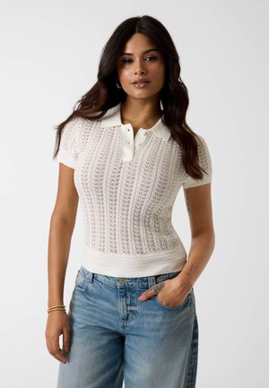 Frau mit langem dunklem Haar trägt ein weißes kurzärmeliges Strick-Poloshirt und blaue Jeans, steht mit einer Hand in der Tasche vor schlichtem Hintergrund.