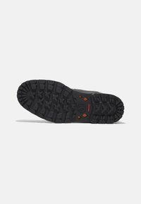 Suela de zapato de goma negra con profundos surcos, que presenta un logo central y acentos en naranja. Superficie texturizada para un mejor agarre y durabilidad.