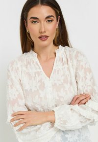 Camicia bianca trasparente con motivo floreale testurizzato, scollatura a volant e chiusura con bottoni. Maniche a palloncino con polsini elasticizzati. Tessuto morbido e leggero.