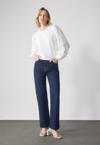 Felpa bianca con maniche a sbuffo in tessuto texture e motivo a occhio di pernice, abbinata a jeans in denim blu scuro a vita alta e sandali bianchi con tacco quadrato.