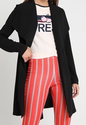 Sort åben cardigan med draperet krave, båret over en lys skjorte med grafisk tryk, parret med røde og hvide stribede leggings.