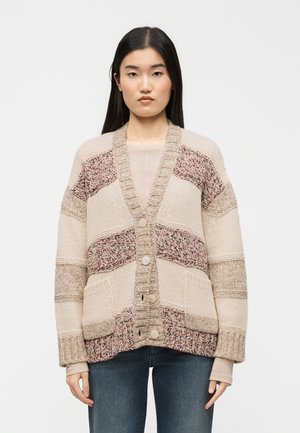 Femme aux longs cheveux bruns portant un cardigan en maille épaisse beige et rose à motifs, et un jean bleu, face à l'avant avec une expression neutre.