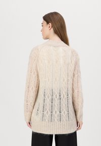 Cardigan tricotat într-o nuanță bej deschis, moale, cu un model de dantelă. Are o croială lungă, lejeră, cu tiv ondulat. Textura pare pufoasă și ușoară.