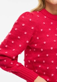 Pull en maille rouge avec des pois roses en relief, manches longues côtelées et encolure ronde porté par une personne aux cheveux blonds.