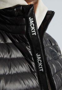 Černá prošívaná bunda s vysokým límcem, viditelným švem a zipem s logem "JACKIT". Lehká a lesklá textura.