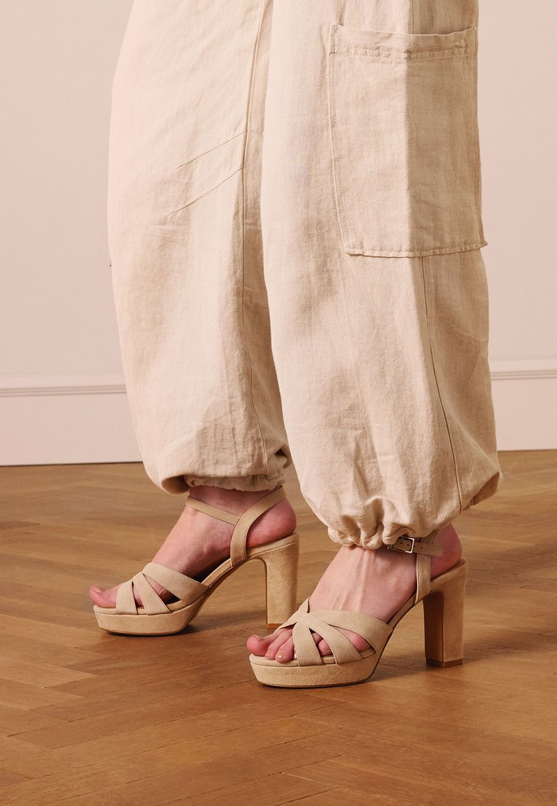 Beige Sandalen mit Absatz und Knöchelriemen, getragen mit lockeren, hellen Leinenhosen mit elastischen Bündchen und Seitentaschen, stehend auf Holzboden.