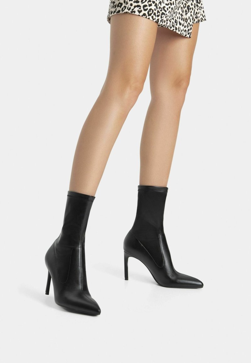 Svarta läderankellågsstövlar med spetsig tå, hög stilettklack och åtsittande ovandel, med en slät textur och elastisk detalj.